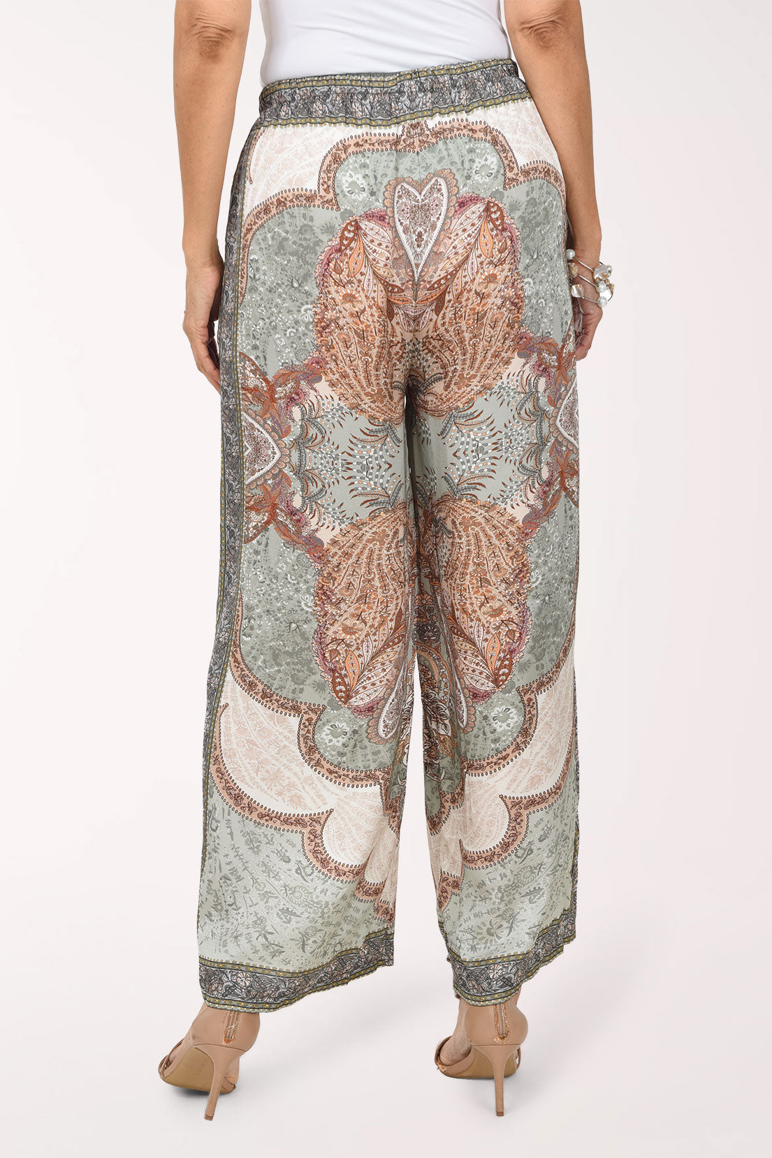 A woman modeling wide-leg patterned pants with a paisley design in muted green, brown, and cream, set against a plain white studio background.