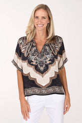 A smiling blonde woman wearing a short-sleeve V-neck top with a black, beige, and brown paisley pattern, paired with white pants, against a plain white studio background.