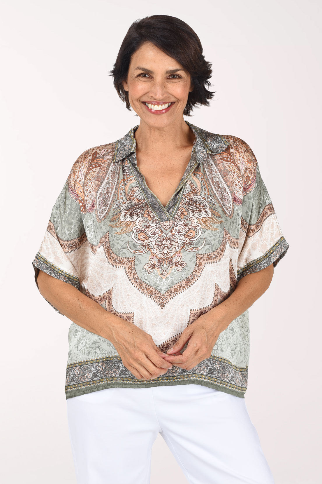 A smiling woman wearing a short-sleeve V-neck blouse with an intricate paisley and floral pattern in beige, sage green, and brown, paired with white pants, against a plain light background.