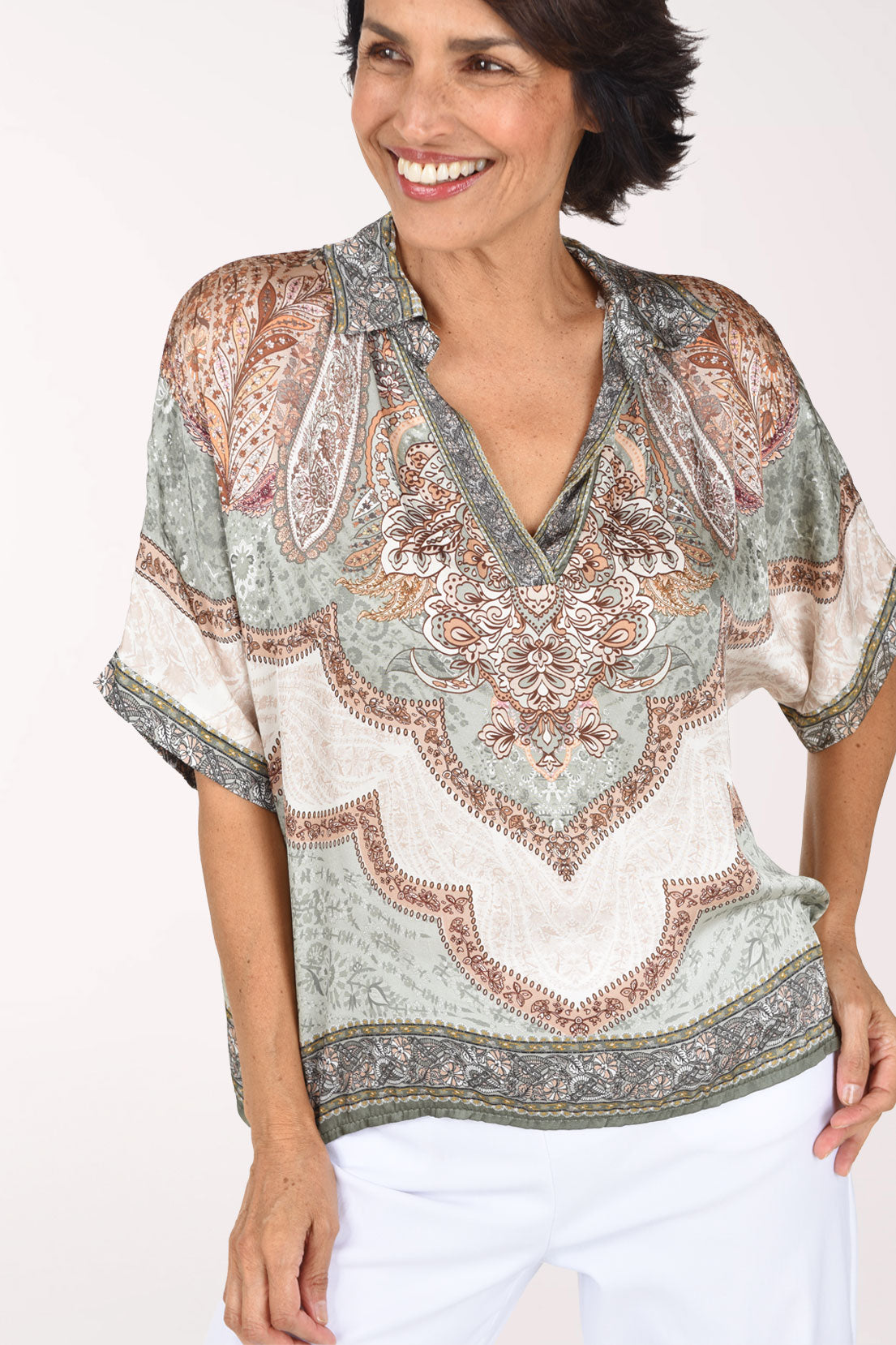 A smiling woman wearing a short-sleeve V-neck blouse with an intricate paisley and floral pattern in beige, sage green, and brown, paired with white pants, against a plain light background.