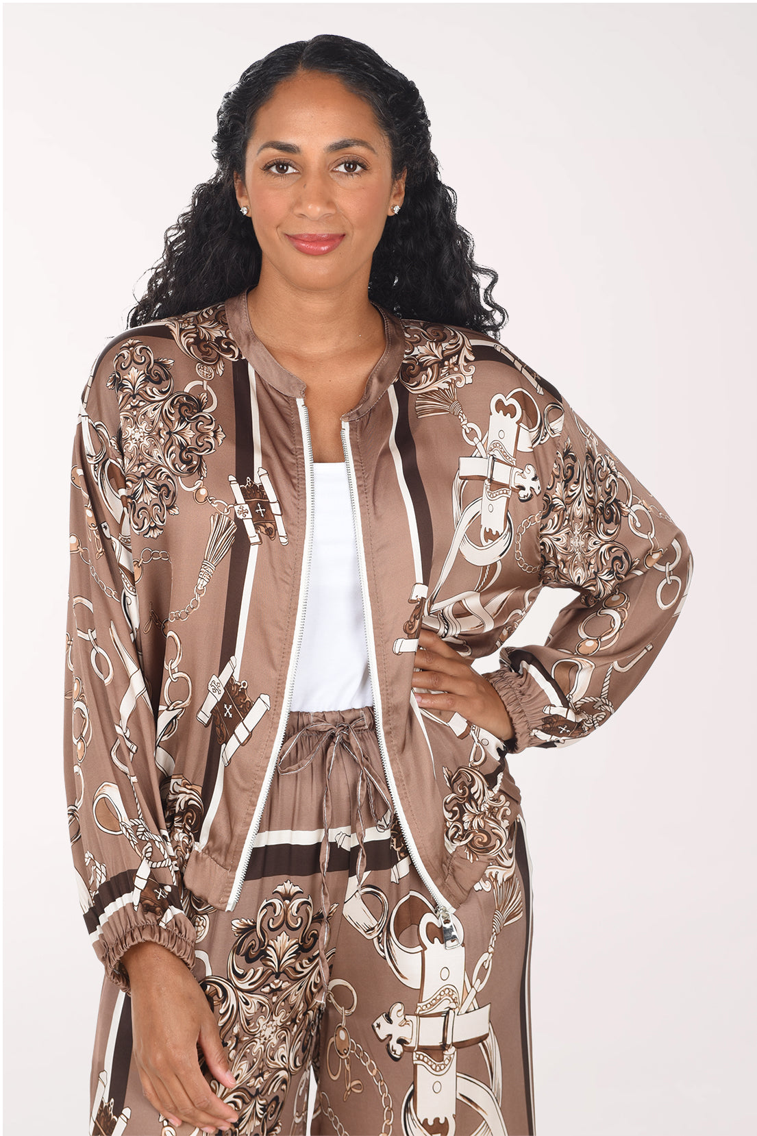 A woman modeling a brown satin zip-up jacket and matching pants set with a white and brown baroque-style pattern, worn over a white tank top, against a plain light background.