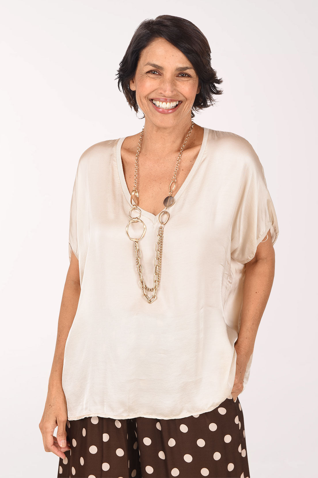 A smiling woman wearing a cream-colored short-sleeve V-neck top with a silky sheen, paired with brown and white polka dot pants and a long gold-toned necklace, set against a plain white studio background.