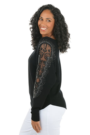 Woman wearing a long sleeve black sweater with scalloped mesh embellishments and rhinestone accents on shoulders. Side shot.