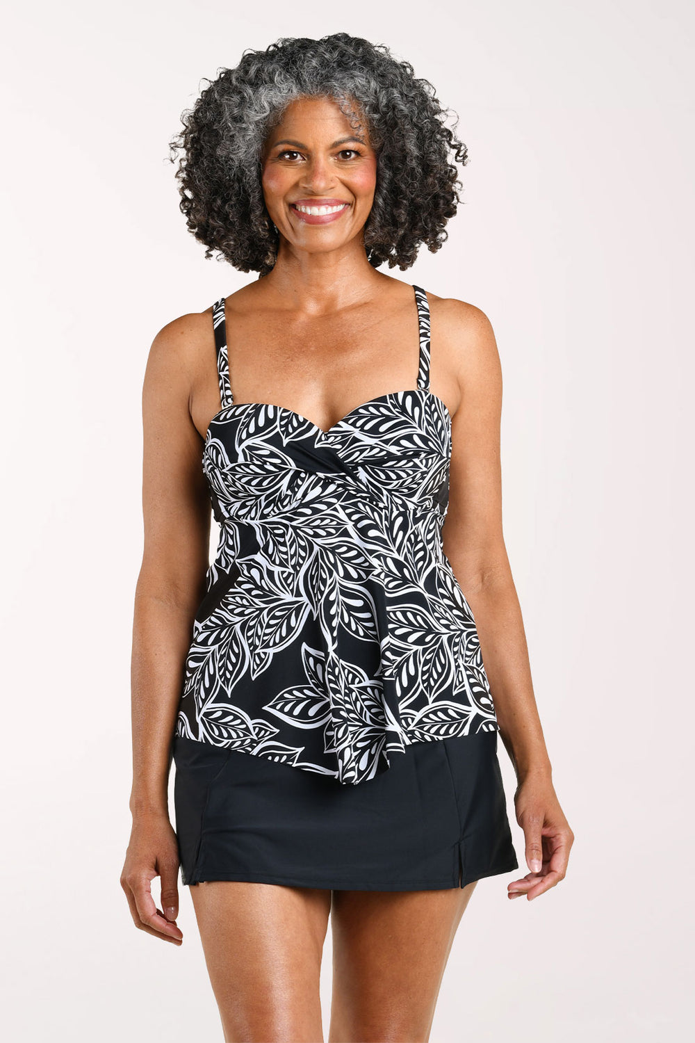 Front view of a woman smiling in the Maxine Native Vines Twisted Bust Tankini Top, a black tankini with a white leaf print, twisted sweetheart bust and flutter hem, worn with a black swim skirt.