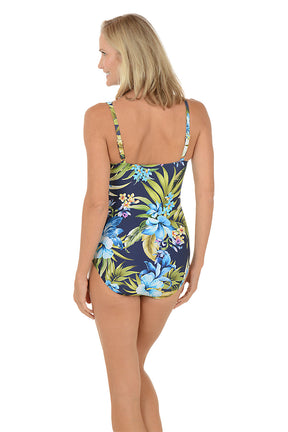 A woman wearing a blue one piece swimsuit with a blue and green floral pattern and a tie on the bust. Back shot.