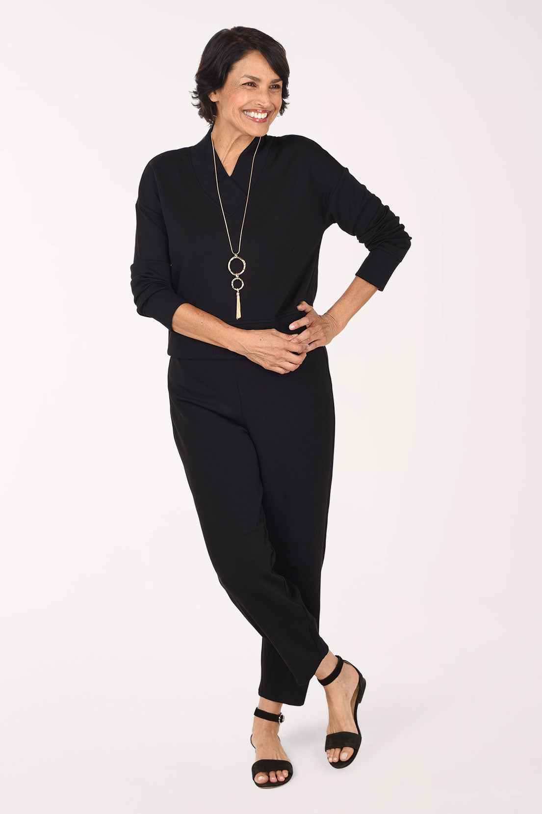 A woman wearing a black long-sleeve top and matching black pants, standing against a plain white studio background.