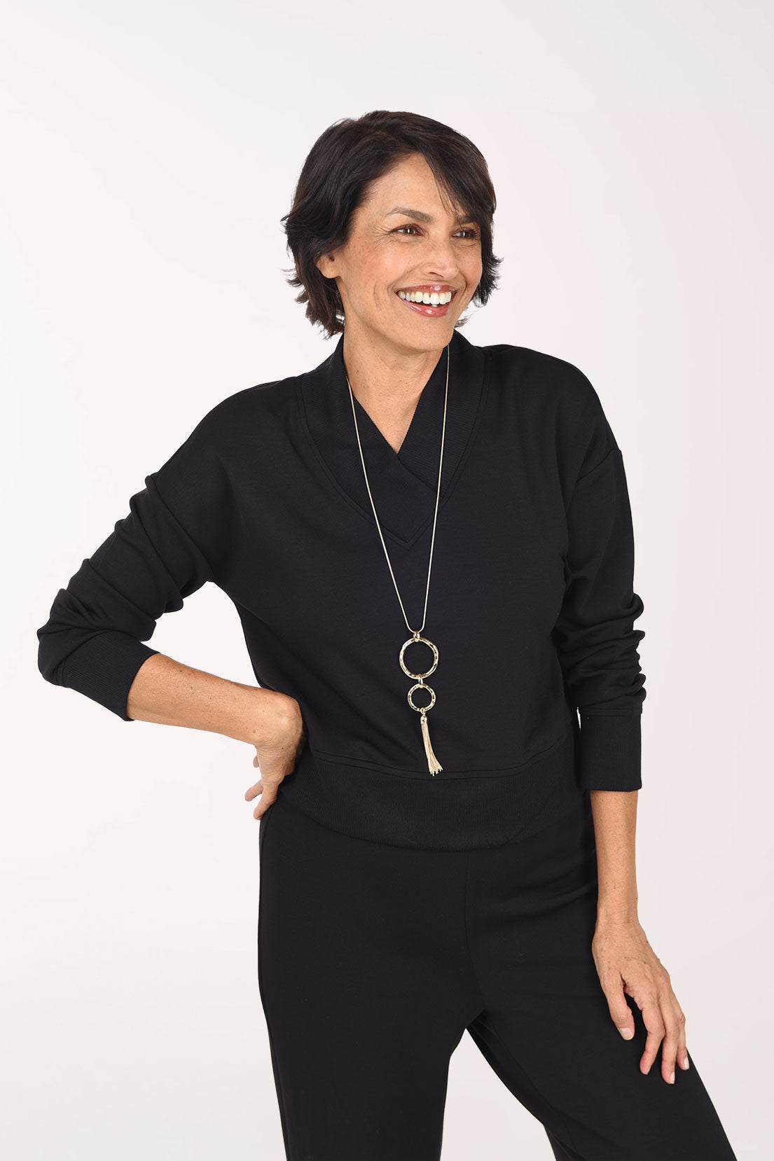 A woman wearing a black long-sleeve top and matching black pants, standing against a plain white studio background.
