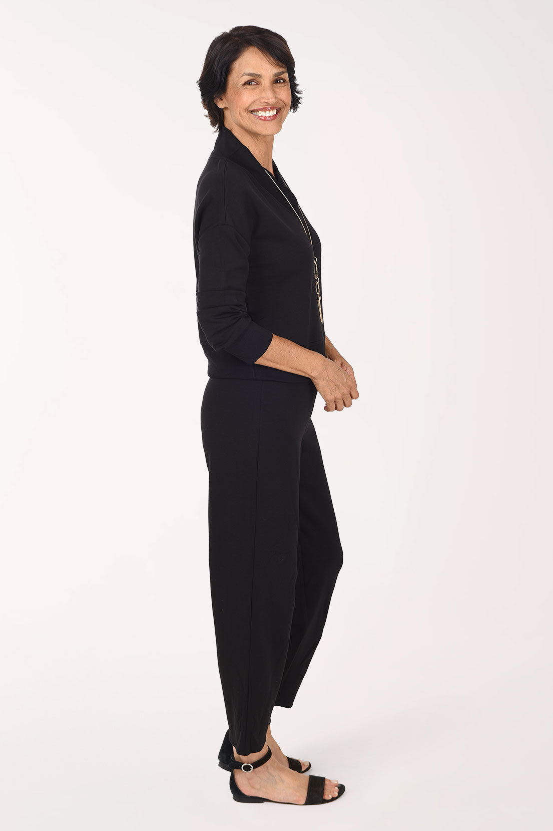 A woman wearing a black long-sleeve top and matching black pants, standing against a plain white studio background.