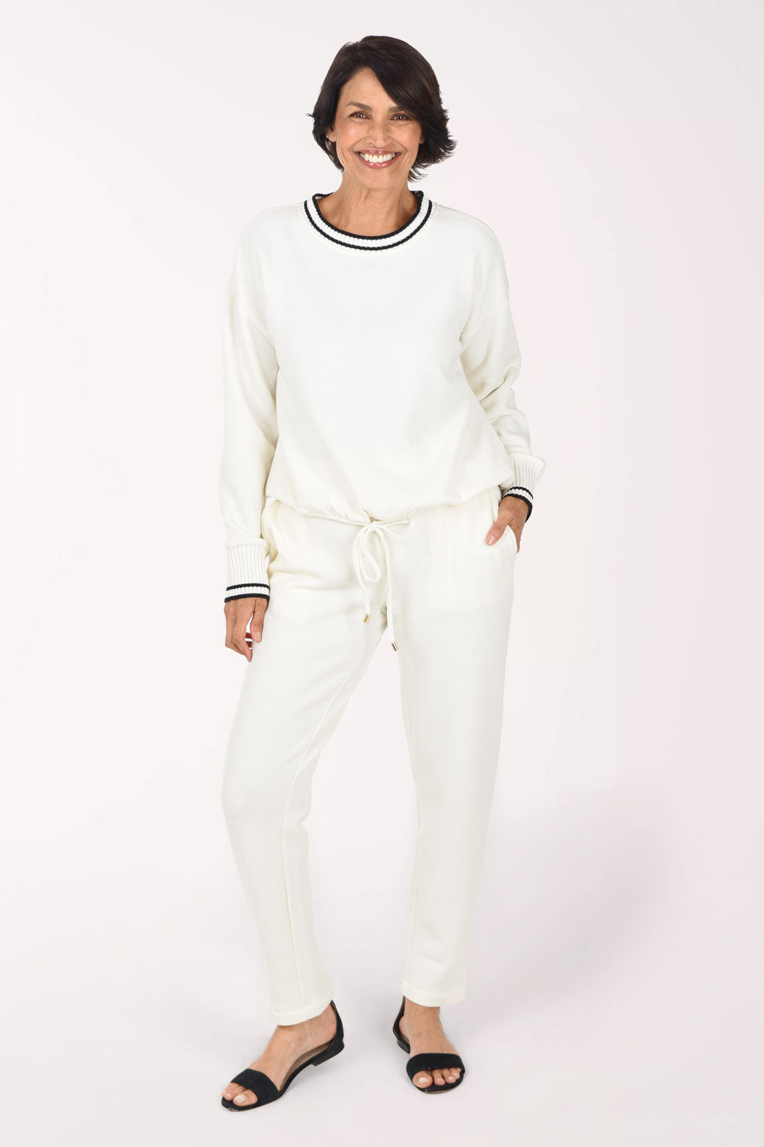 A woman modeling a matching cream lounge set consisting of a long-sleeve crewneck sweatshirt with black and white striped trim and matching drawstring sweatpants, paired with black slide sandals against a light studio background.