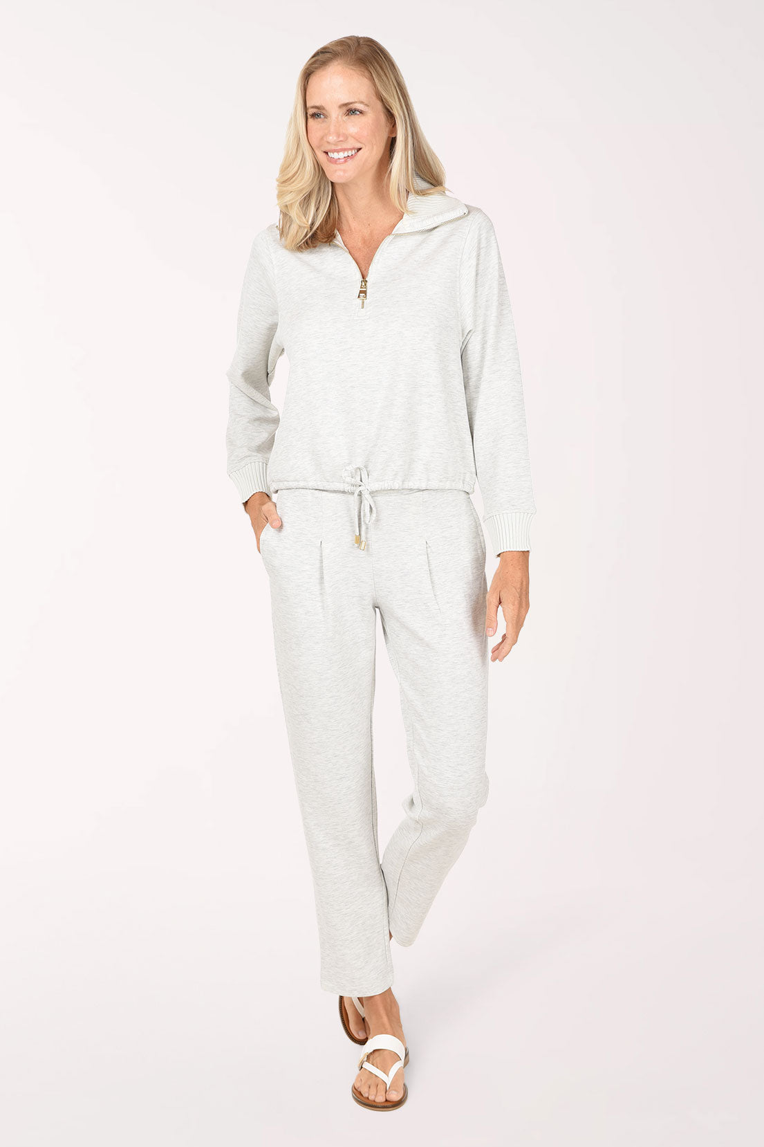 A woman wearing a light grey half-zip sweater and matching grey pants, standing against a plain white studio background.