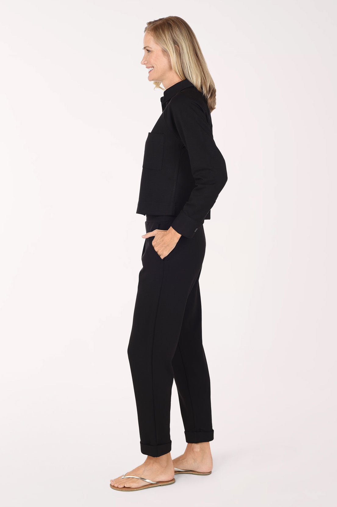 A woman in a side profile wearing a black long-sleeve top and matching black pants, standing against a plain white studio background.