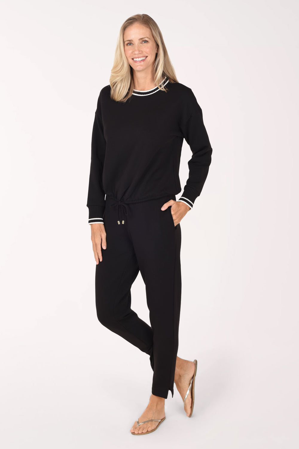 A woman modeling a black two-piece lounge set consisting of a long-sleeve crewneck top with white striped trim and matching black drawstring pants, set against a plain white studio background.