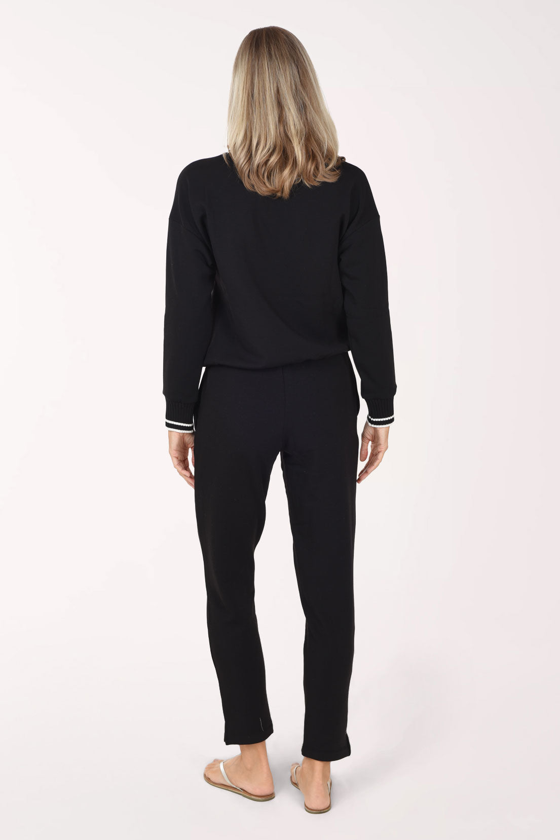 A woman viewed from the back wearing a black long-sleeve top and black pants with white striped cuffs, standing against a plain light background.