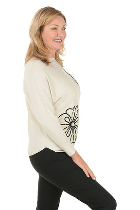 Woman wearing a cream sweater with black floral designs on a white background. Side shot.
