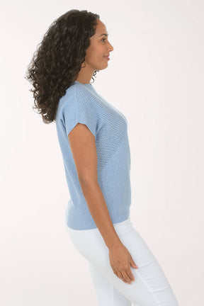 Woman wearing a blue textured top and white pants on a light background