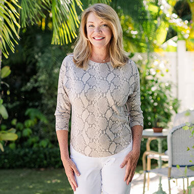 Woman wearing a patterned top and white pants standing outdoors with greenery in the background