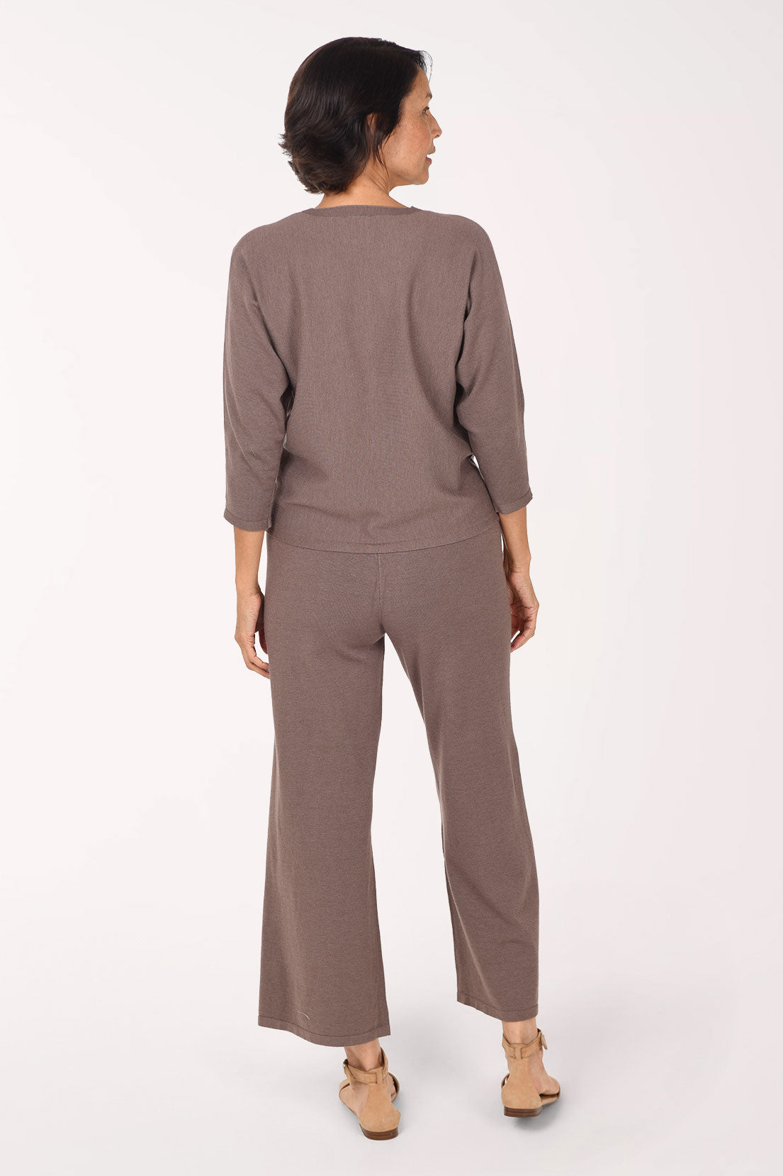 A woman modeling a taupe knit matching set with three-quarter sleeves and wide legs, paired with beige heeled sandals, against a plain white studio background.