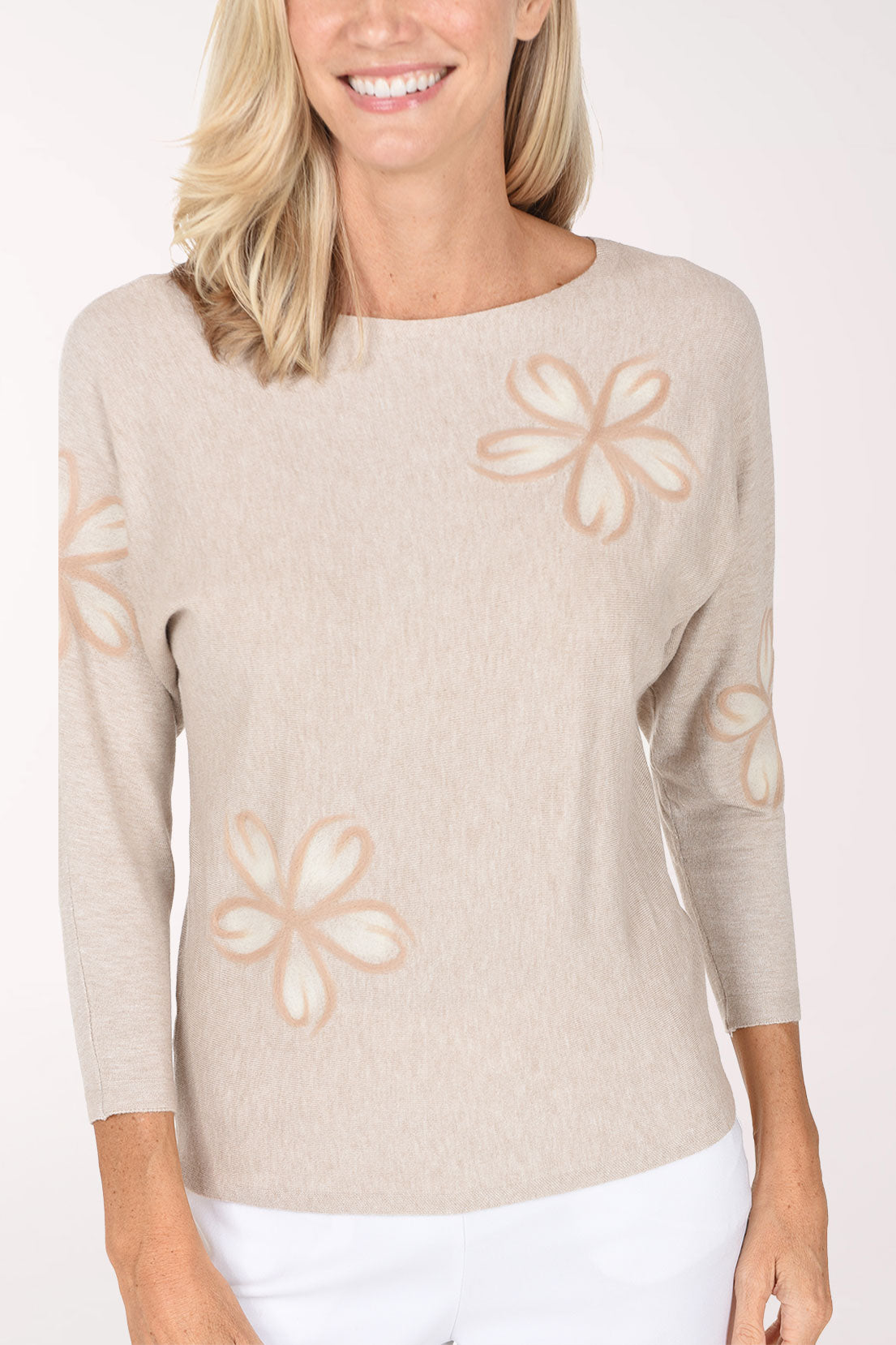 A woman wearing a light beige knit sweater with a tonal embroidered flower details, paired with white pants, against a plain white studio background.