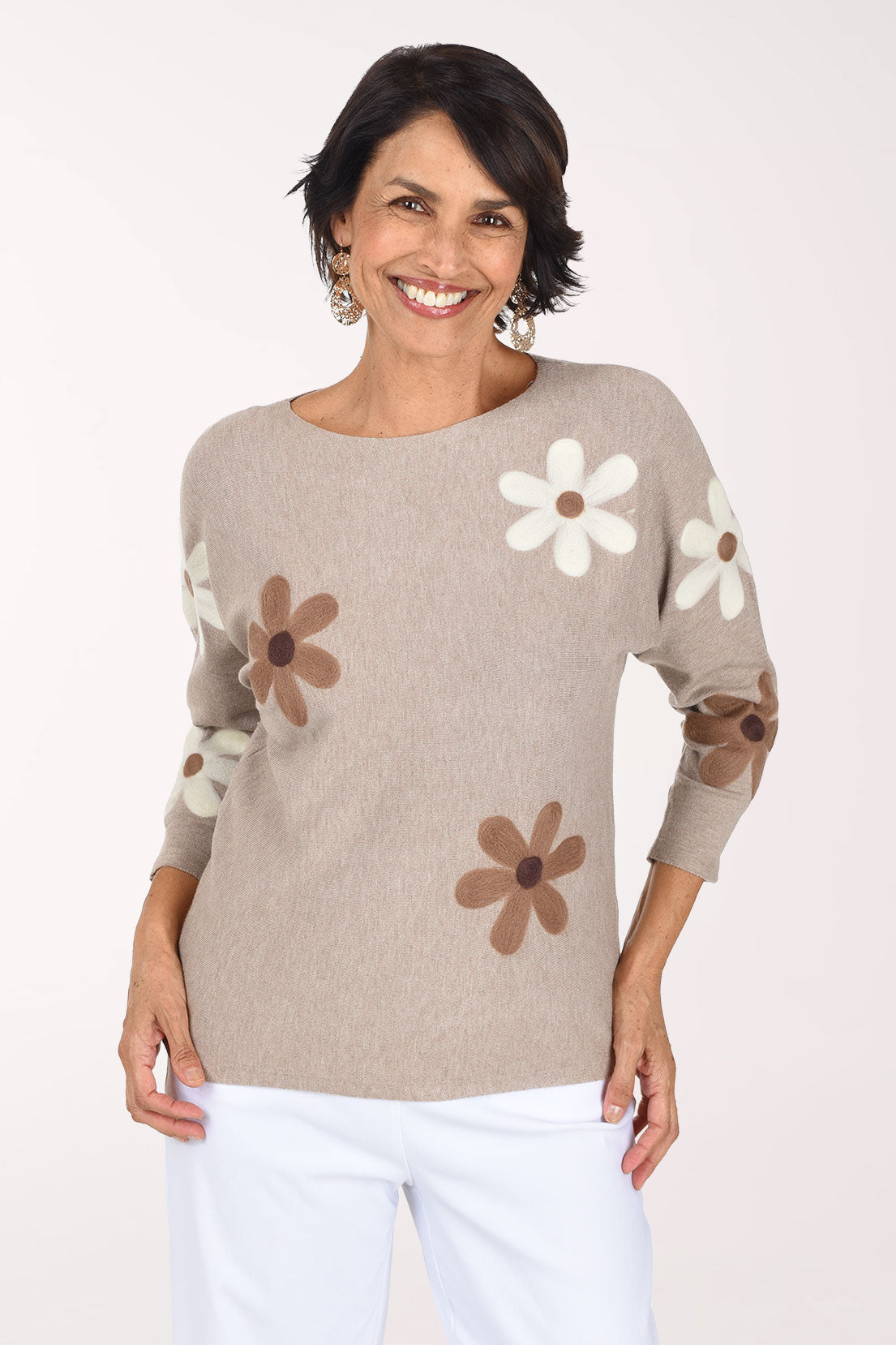 A smiling woman wearing a beige knit sweater with large brown and white daisy patches on the sleeves and body, paired with white pants, against a plain white studio background.