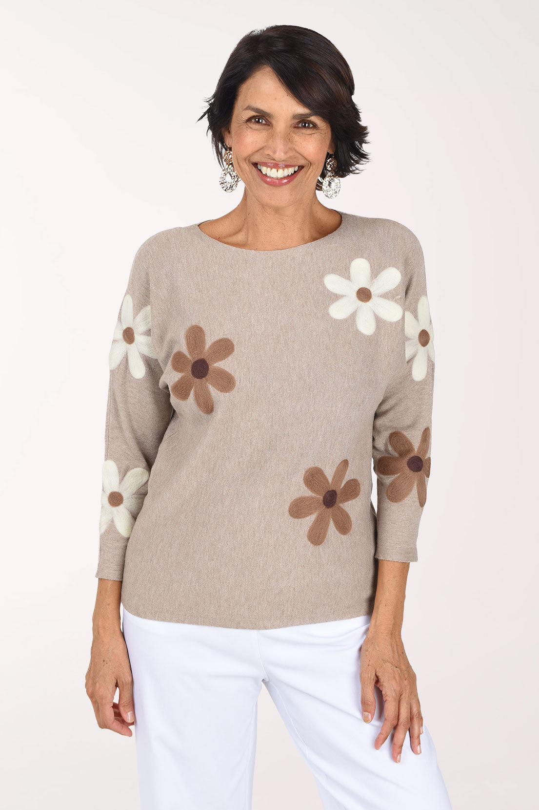 A smiling woman wearing a beige knit sweater with large brown and white daisy patches on the sleeves and body, paired with white pants, against a plain white studio background.