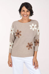 A smiling woman wearing a beige knit sweater with large brown and white daisy patches on the sleeves and body, paired with white pants, against a plain white studio background.