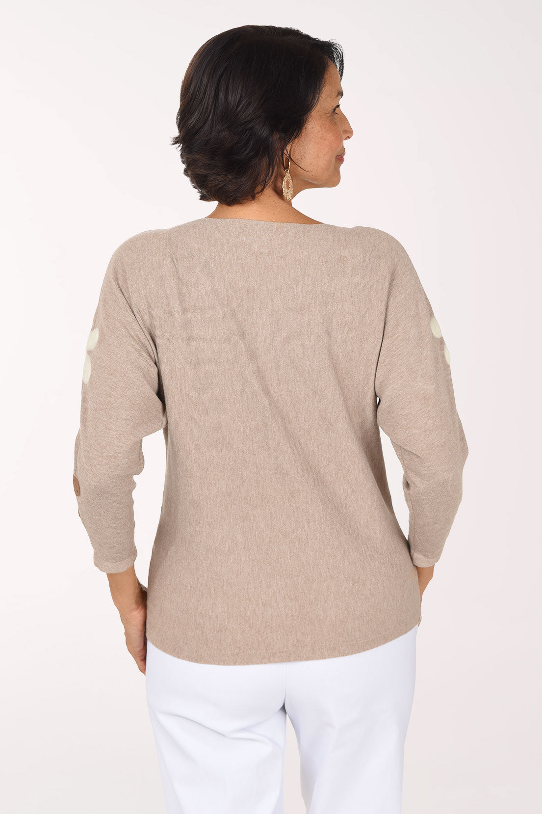 A smiling woman wearing a beige knit sweater with large brown and white daisy patches on the sleeves and body, paired with white pants, against a plain white studio background.