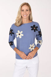 A smiling blonde woman wearing a dusty blue knit sweater with large black and white daisy patches on the sleeves and body, paired with white pants, against a plain white studio background.