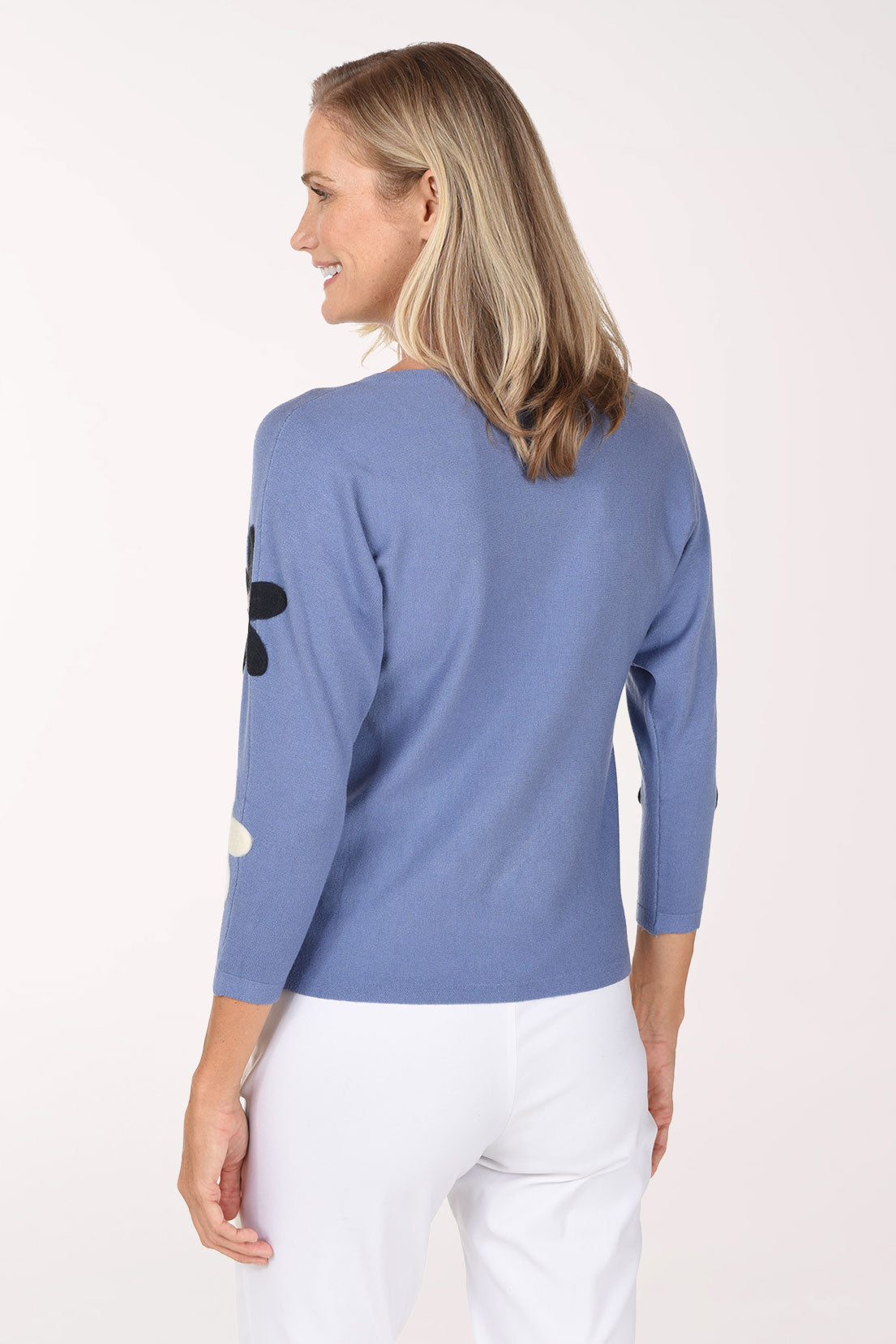A smiling blonde woman wearing a dusty blue knit sweater with large black and white daisy patches on the sleeves and body, paired with white pants, against a plain white studio background.