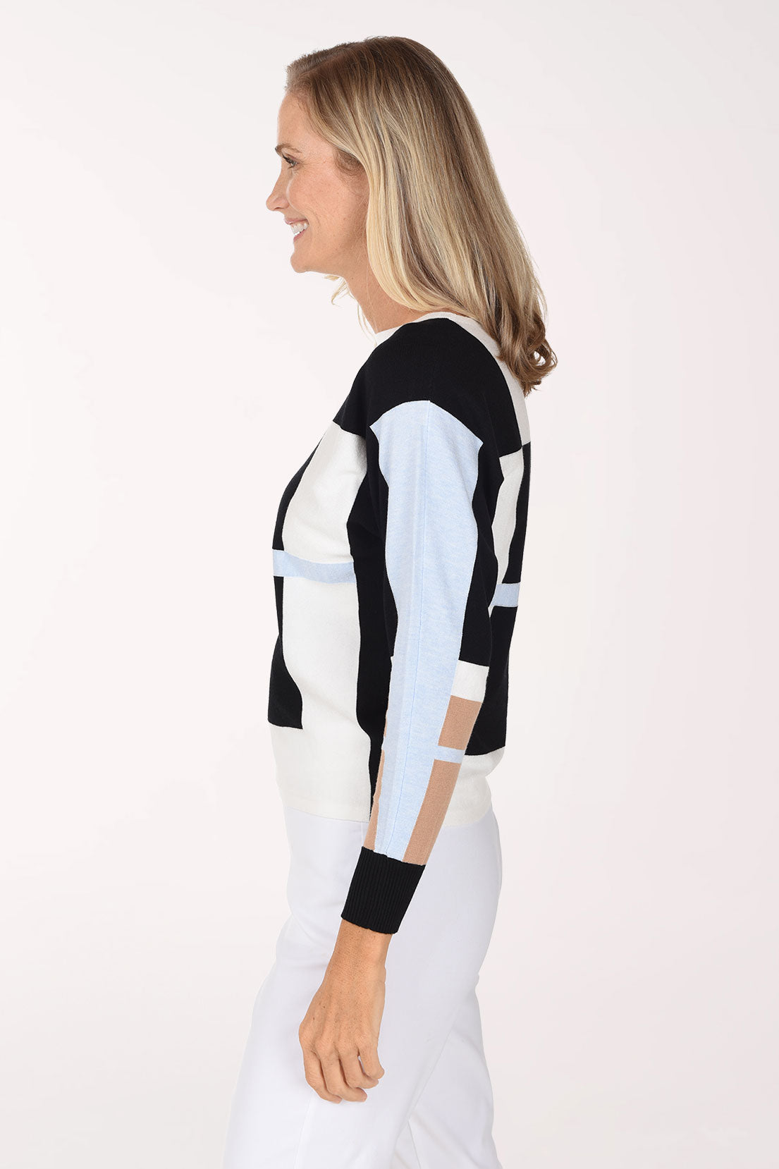 A woman modeling a color-block knit sweater with black, white, light blue, and beige sections, paired with white pants, against a plain light background.