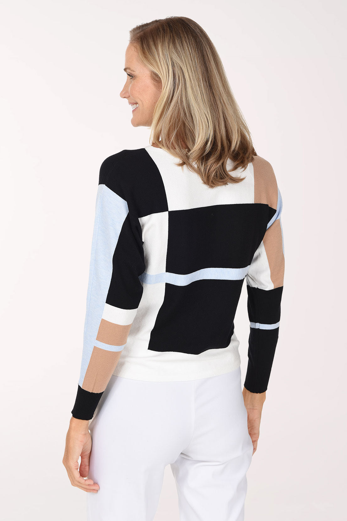 A woman modeling a color-block knit sweater with black, white, light blue, and beige sections, paired with white pants, against a plain light background.