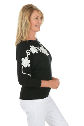 Woman wearing a black sweater with white floral patterned embroidery on a white background. Side shot.