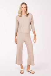 A woman modeling a matching two-piece knit loungewear set in a light taupe-beige color, featuring a three-quarter sleeve top and wide-leg cropped pants, paired with white slide sandals against a plain white studio background.