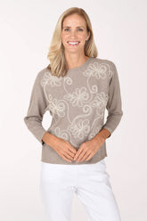 Woman wearing a beige 3/4 sleeve sweater with a white pearl flower design 