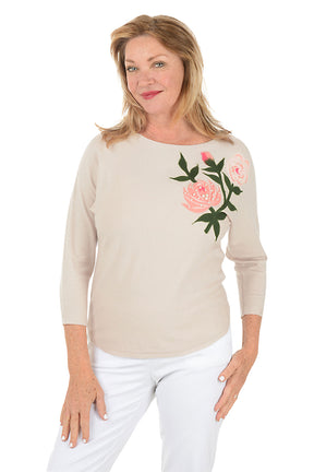 Woman wearing a beige sweater with floral embroidery on a white background.