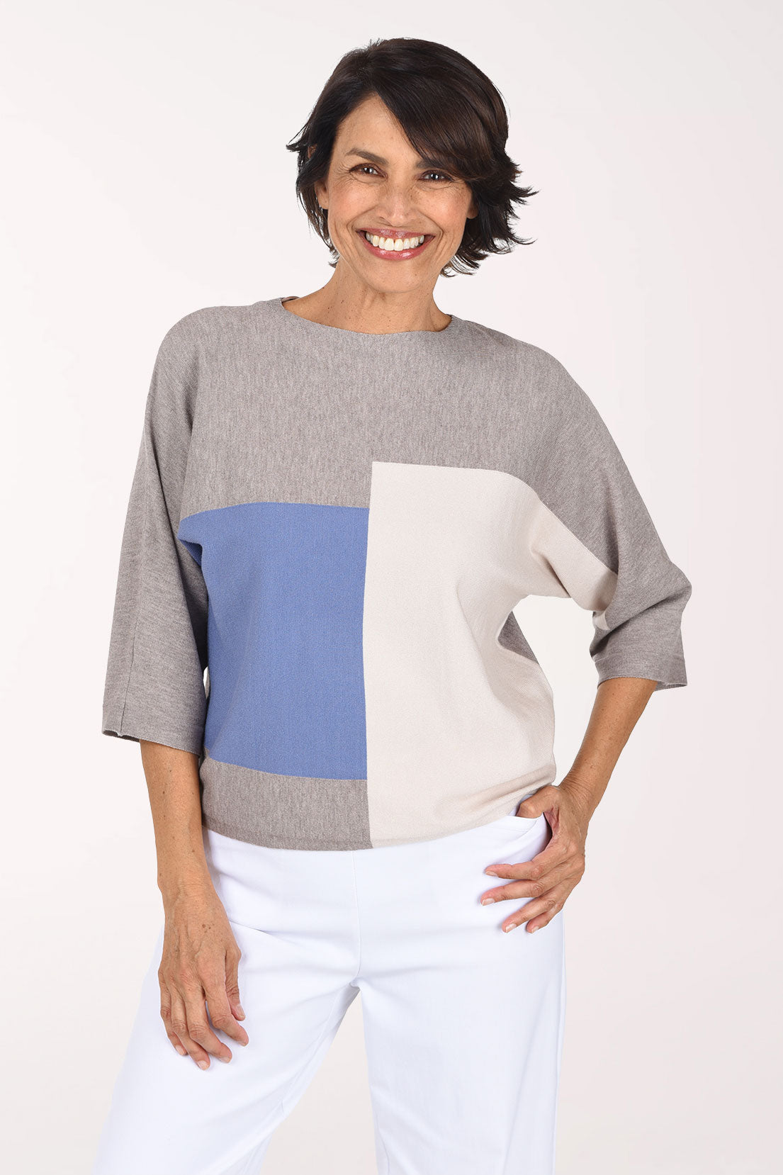 A woman modeling a color-block knit sweater with three-quarter sleeves, featuring a heather gray body, a light blue front panel, and a cream side panel, paired with white pants against a plain white studio background.