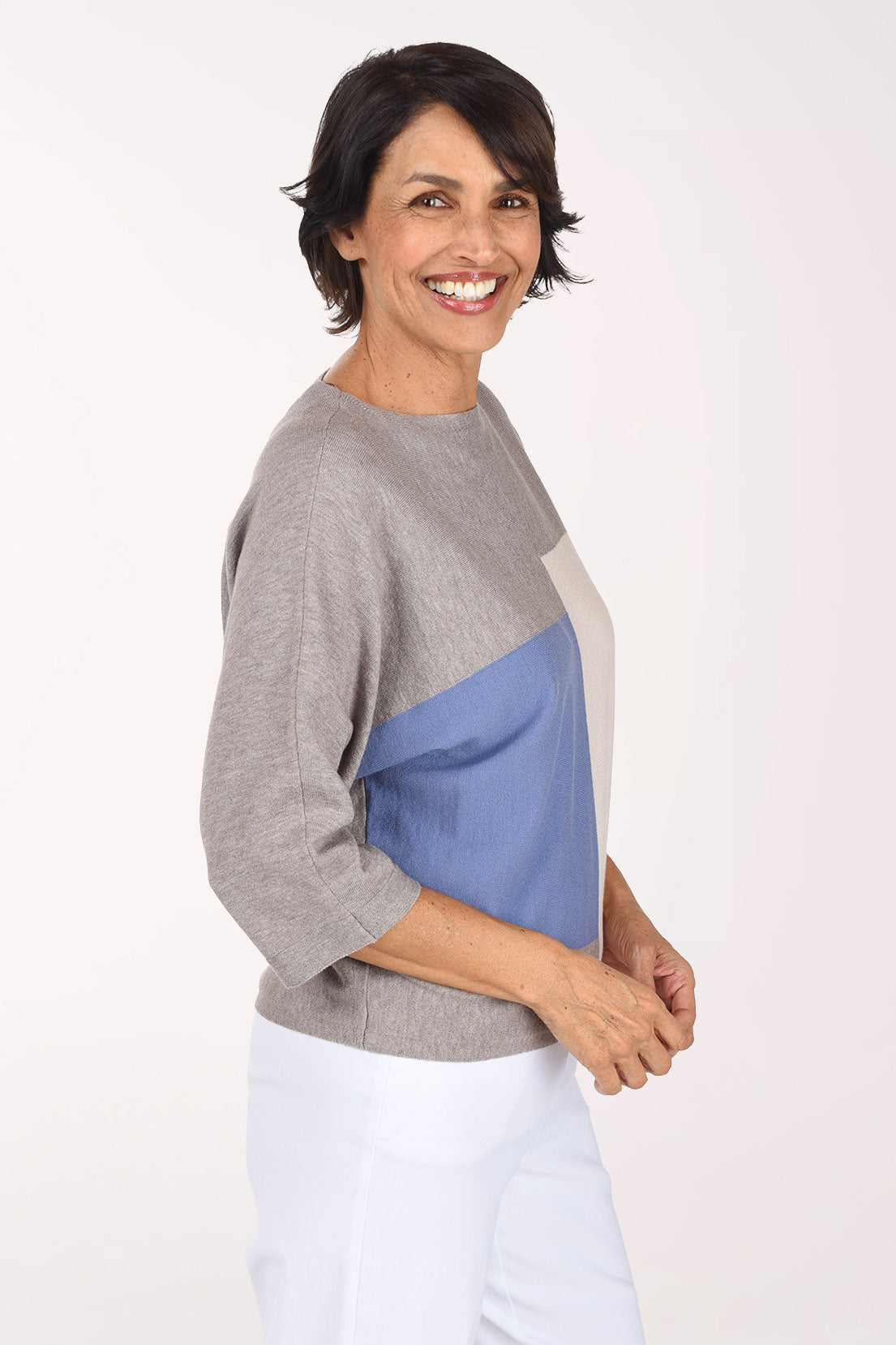 A woman modeling a color-block knit sweater with three-quarter sleeves, featuring a heather gray body, a light blue front panel, and a cream side panel, paired with white pants against a plain white studio background.
