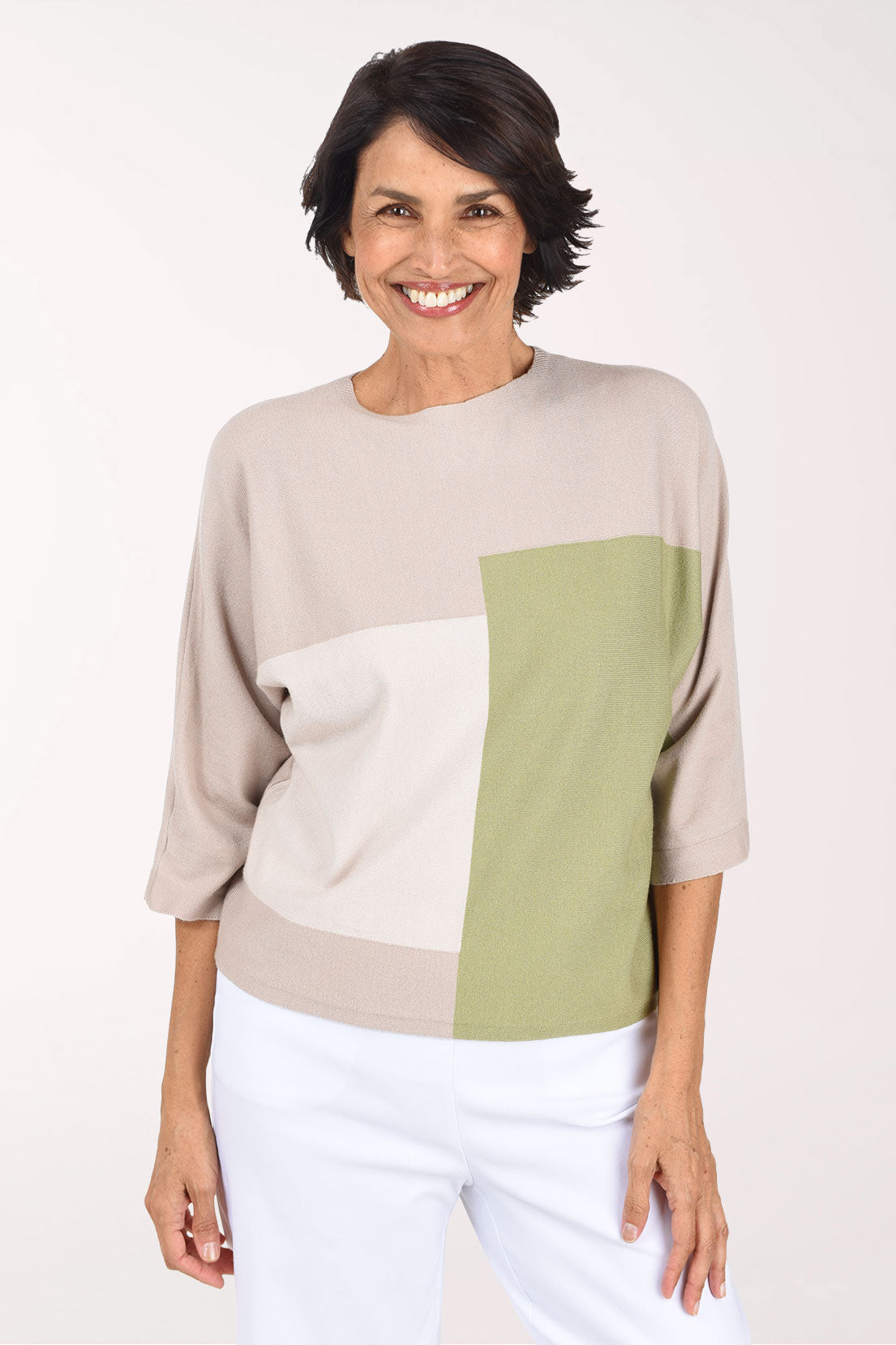 A smiling woman wearing a color-block knit sweater with beige, white, and green panels, paired with white pants, against a plain white studio background.