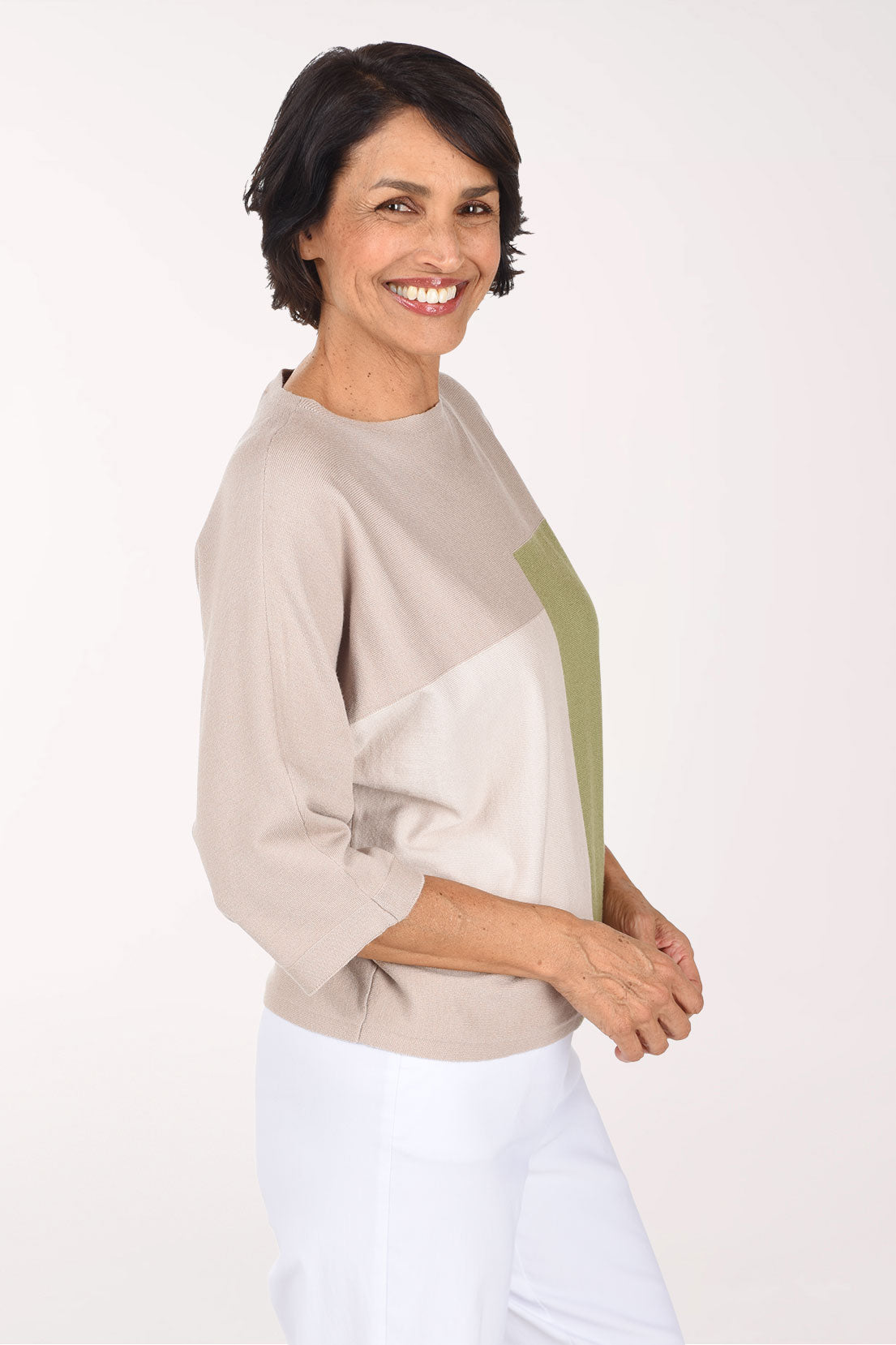 A smiling woman wearing a color-block knit sweater with beige, white, and green panels, paired with white pants, against a plain white studio background.