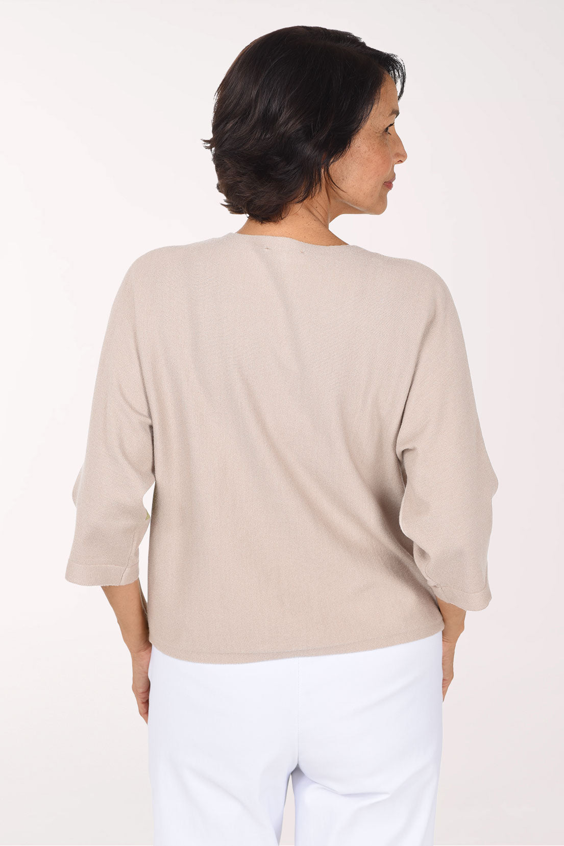 A smiling woman wearing a color-block knit sweater with beige, white, and green panels, paired with white pants, against a plain white studio background.