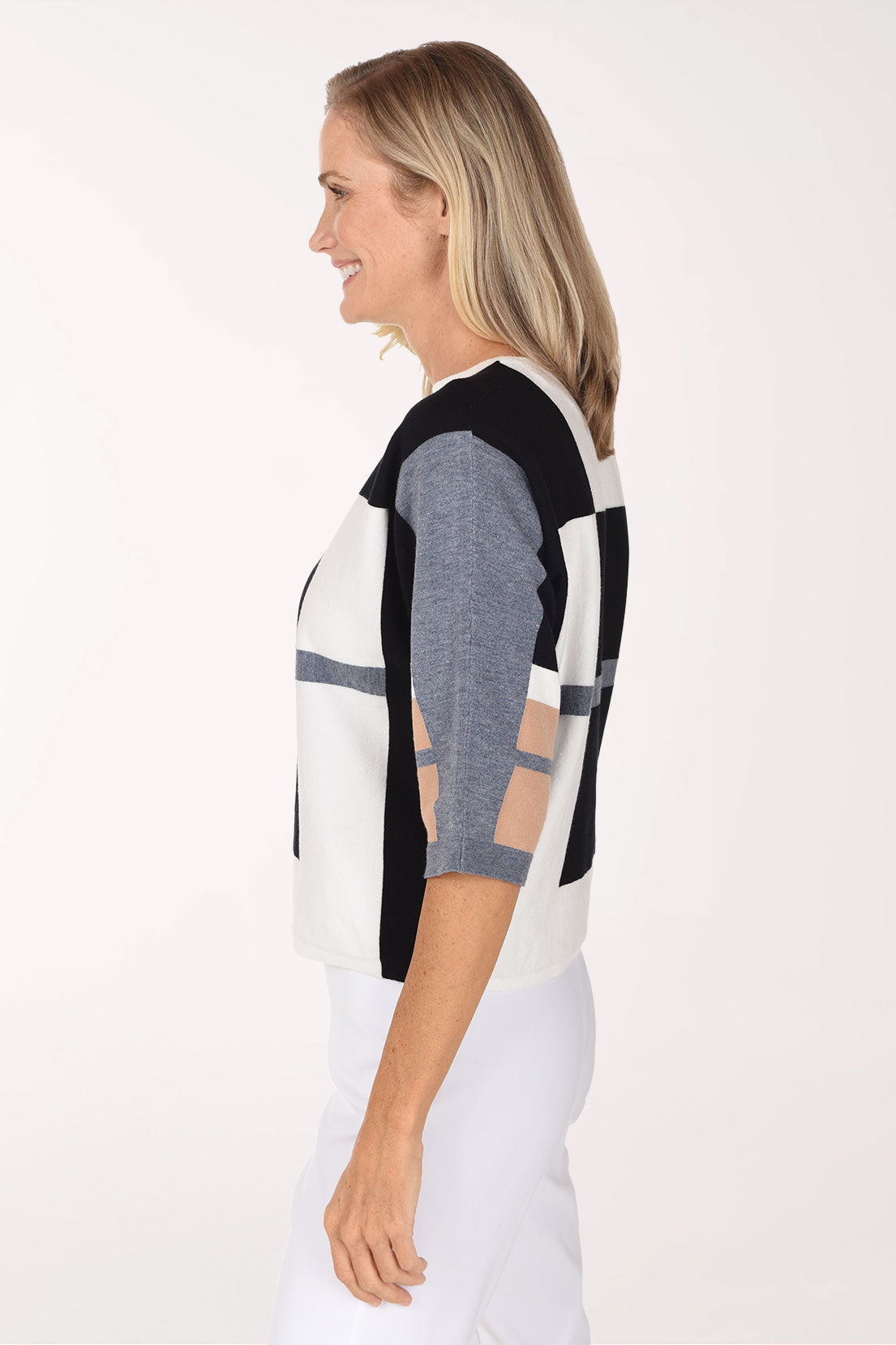 A smiling blonde woman wearing a color-block knit sweater with black, white, gray, and beige panels, paired with white pants, against a plain white studio background.