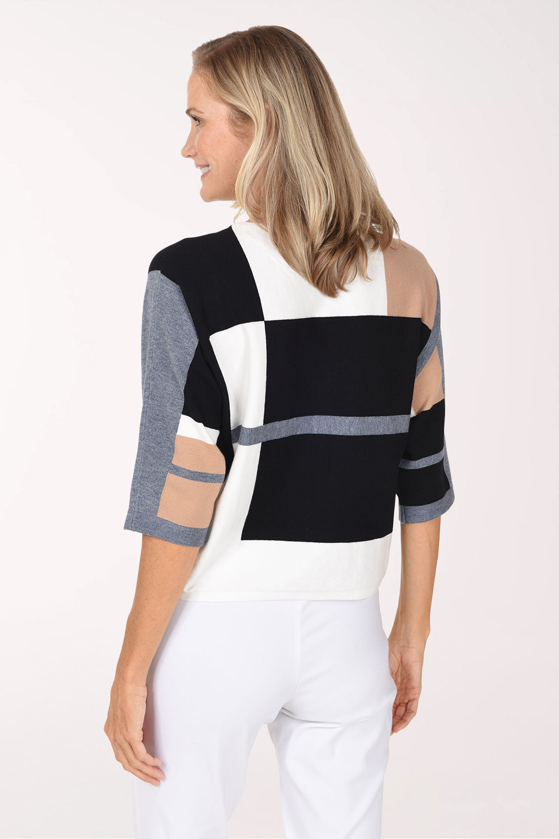 A smiling blonde woman wearing a color-block knit sweater with black, white, gray, and beige panels, paired with white pants, against a plain white studio background.