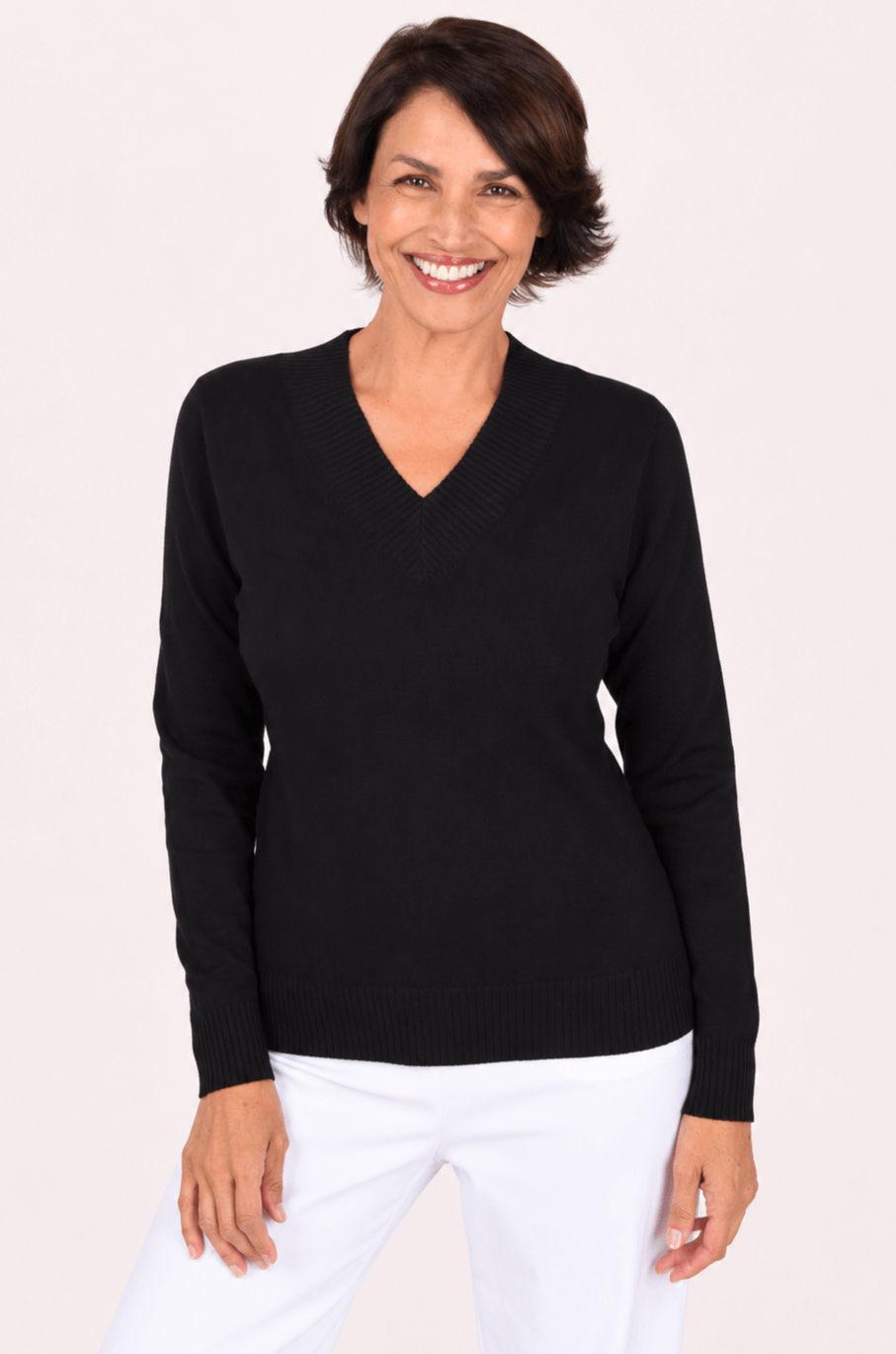 A smiling woman with short brown hair wearing a black long-sleeve V-neck knit sweater and white pants, standing against a plain light background.