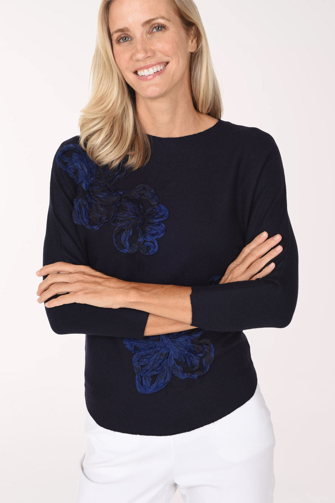 Woman wearing a 3/4 sleeve black sweater with a blue ribbon floral embroidered detail on shoulder and bottom of sweater 