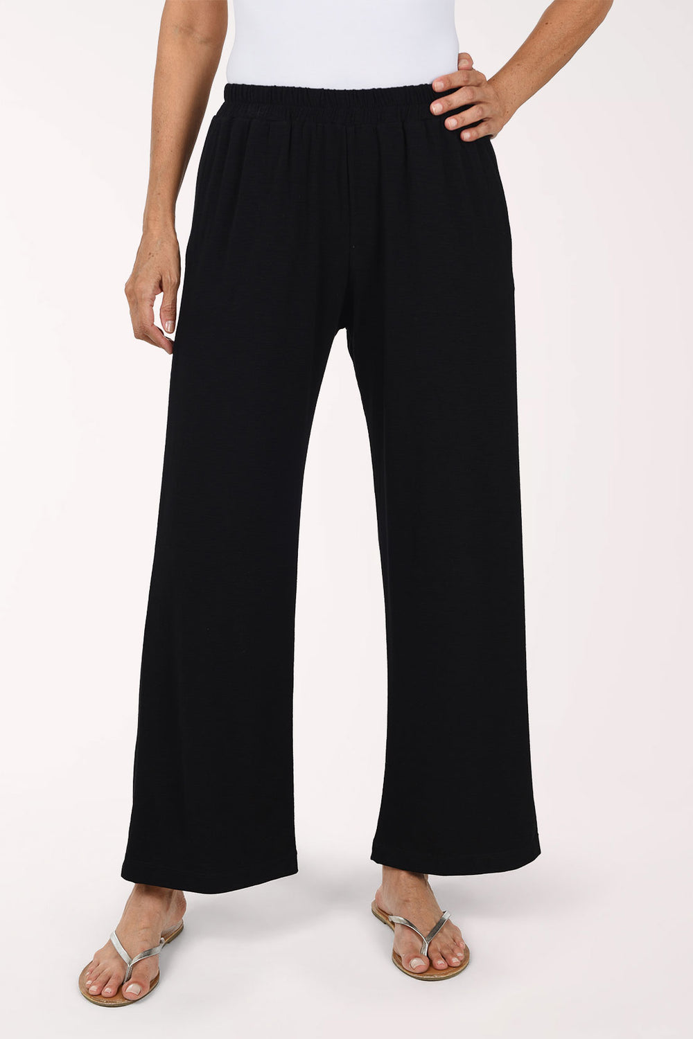A person wearing black  wide-leg pants with an elastic waistband and side pockets, paired with a white top and flat sandals, photographed against a plain light background.
