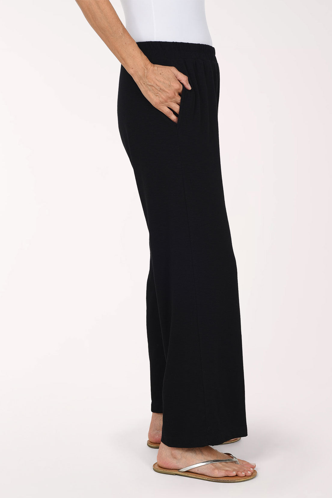 A person wearing black  wide-leg pants with an elastic waistband and side pockets, paired with a white top and flat sandals, photographed against a plain light background.