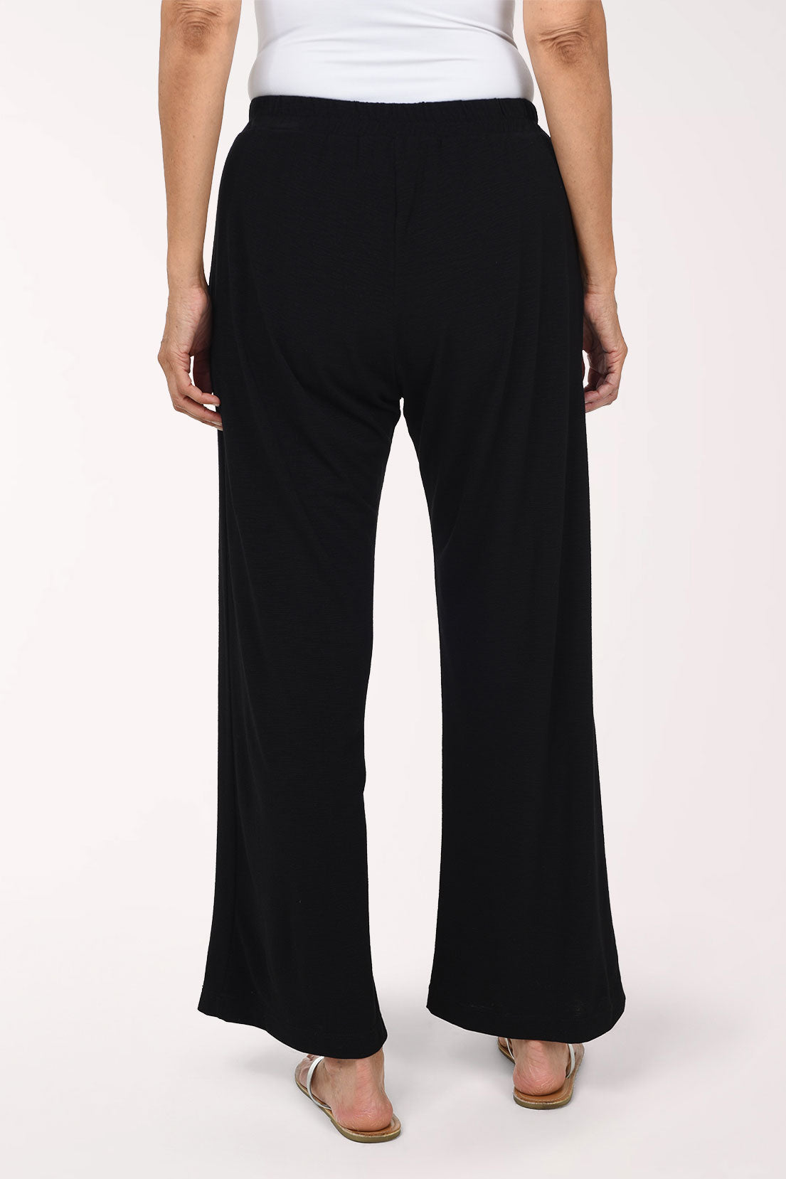 A person wearing black  wide-leg pants with an elastic waistband and side pockets, paired with a white top and flat sandals, photographed against a plain light background.