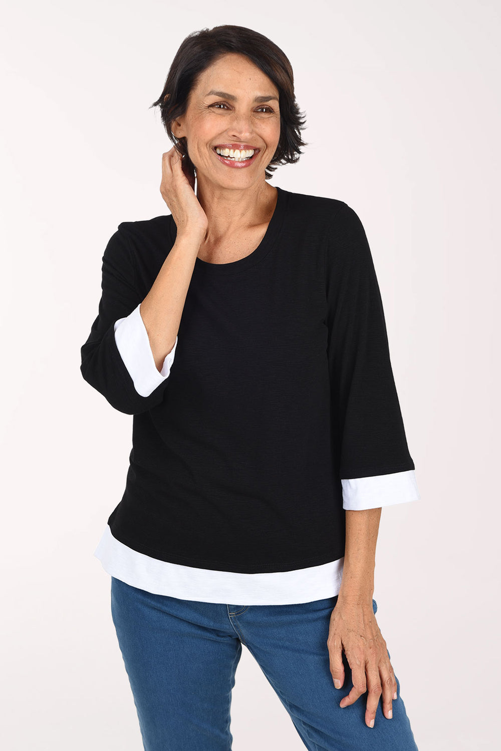 A smiling woman wearing a black three-quarter sleeve top with white contrast trim at the collar, cuffs, and hem, paired with blue jeans, against a plain light background.
