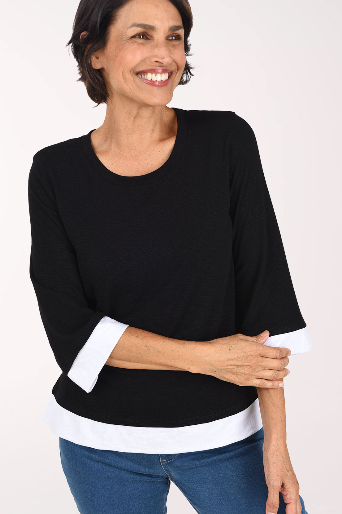A smiling woman wearing a black three-quarter sleeve top with white contrast trim at the collar, cuffs, and hem, paired with blue jeans, against a plain light background.