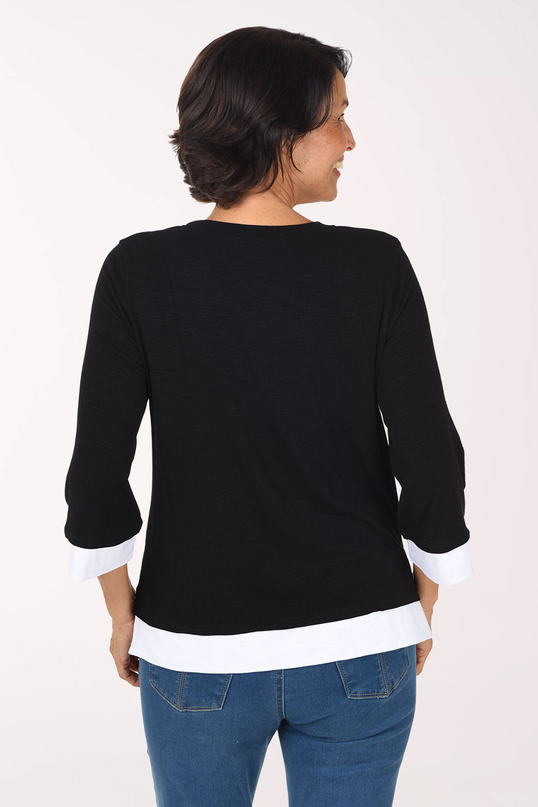 A smiling woman wearing a black three-quarter sleeve top with white contrast trim at the collar, cuffs, and hem, paired with blue jeans, against a plain light background.