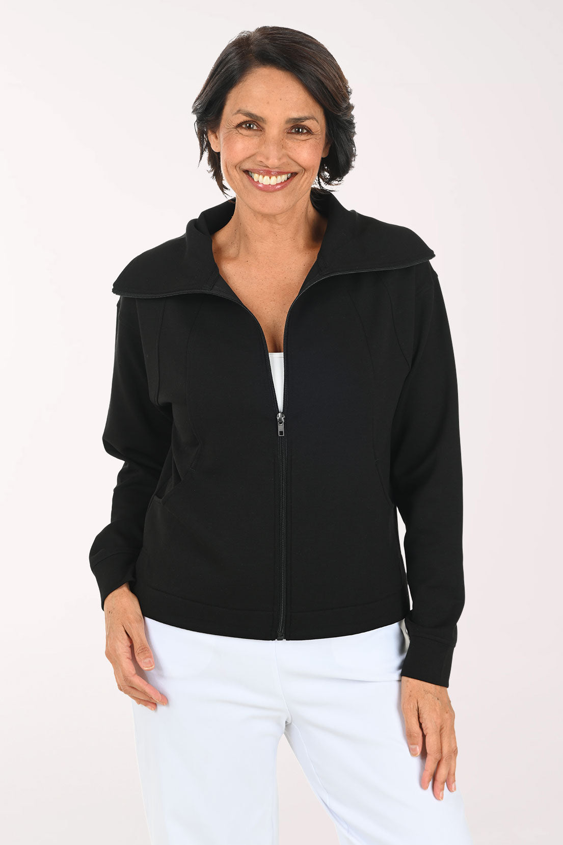 A smiling woman wearing a black zip-up jacket over a white top and white pants, standing against a plain light background.